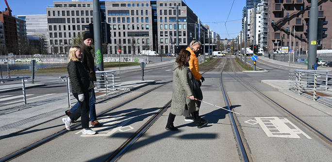 Kvinne med hvit stokk krysser trikkeskinner på fotgjengerfelt sammen med to kvinner og en mann